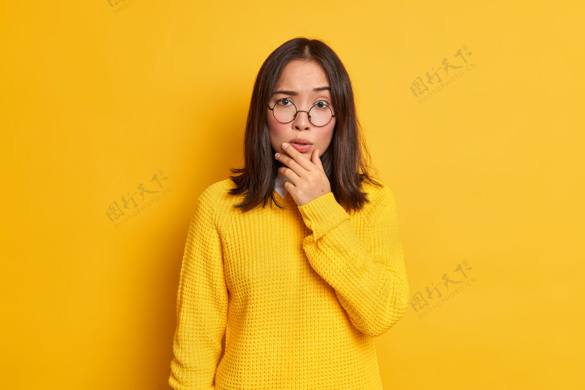 室内忧心忡忡的亚洲女人的画像托着下巴看起来很关心戴着透明眼镜和毛衣站立可爱姿势
