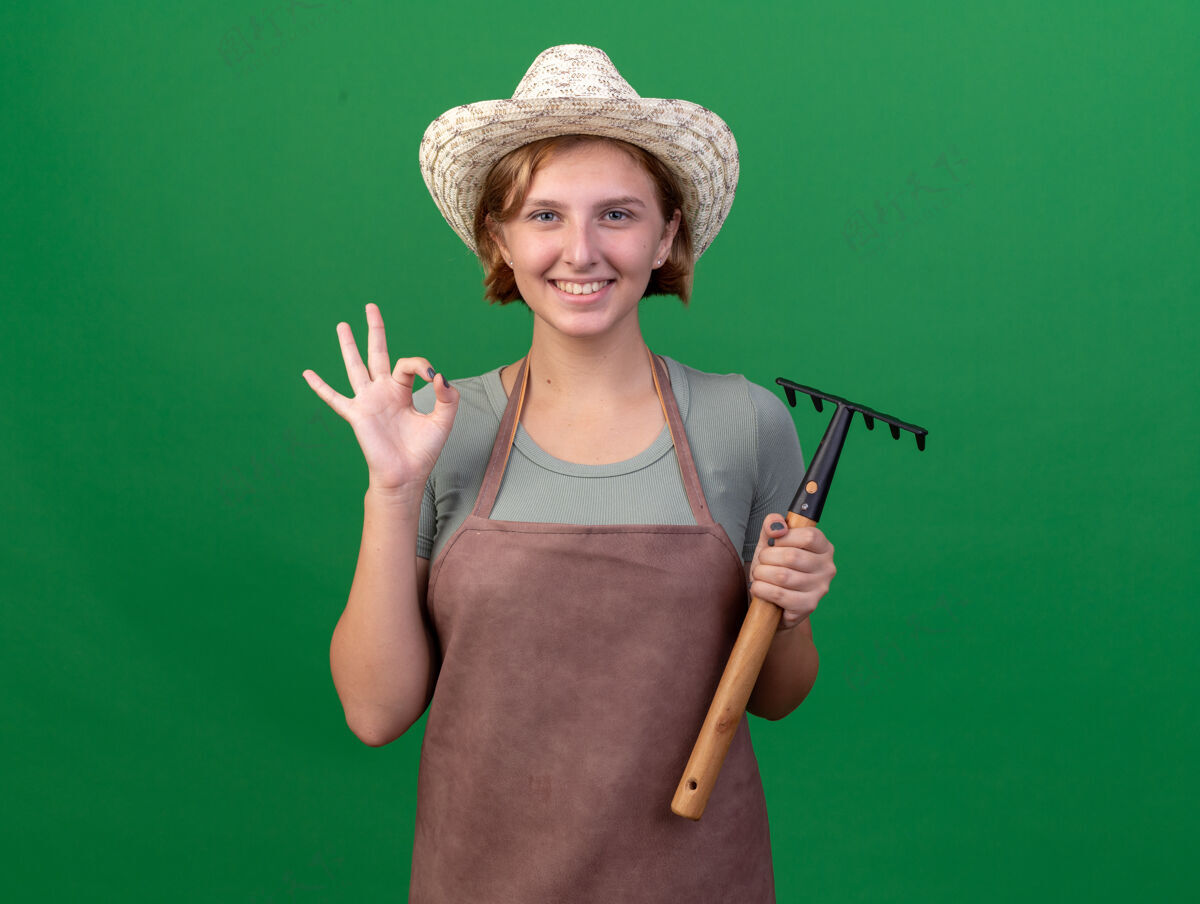 手势面带微笑的年轻斯拉夫女园丁戴着园艺帽 打着“ok”的手势 手里拿着绿色的耙子花园帽子年轻的