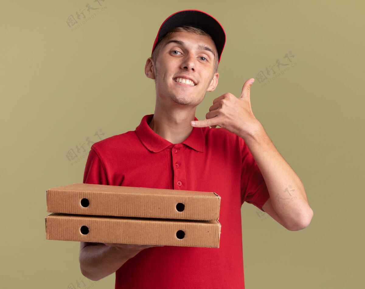 披萨微笑的年轻金发送货男孩拿着比萨饼盒 手势叫我橄榄绿上的标志送货手势微笑