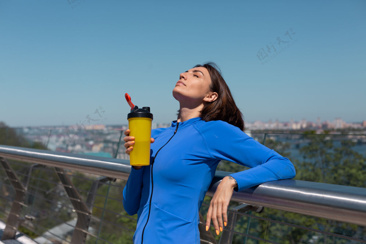 锻炼年轻女子穿着合身的运动服 在炎热阳光明媚的早晨 在桥上喝着一瓶水 运动后口渴又累女性慢跑运动员