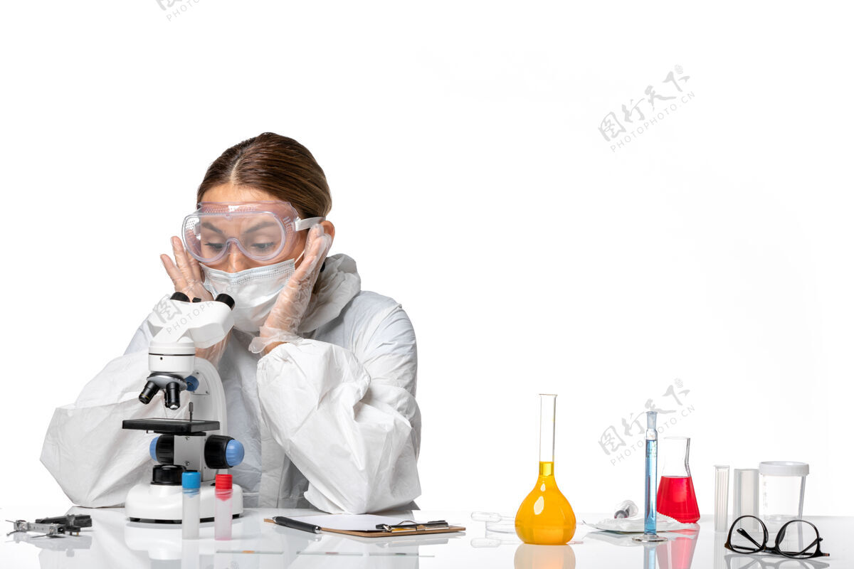 女医生前视女医生穿着特制的工作服 戴着口罩 用显微镜对着白色办公桌上的冠状病毒大流行病毒桌子实验室