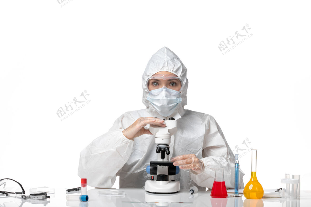 浅白色前视图：女医生穿着防护服 戴着口罩 在显微镜下观察因病毒感染而引起的浅白色背景病毒大流行的健康病毒面罩使用实验室外套