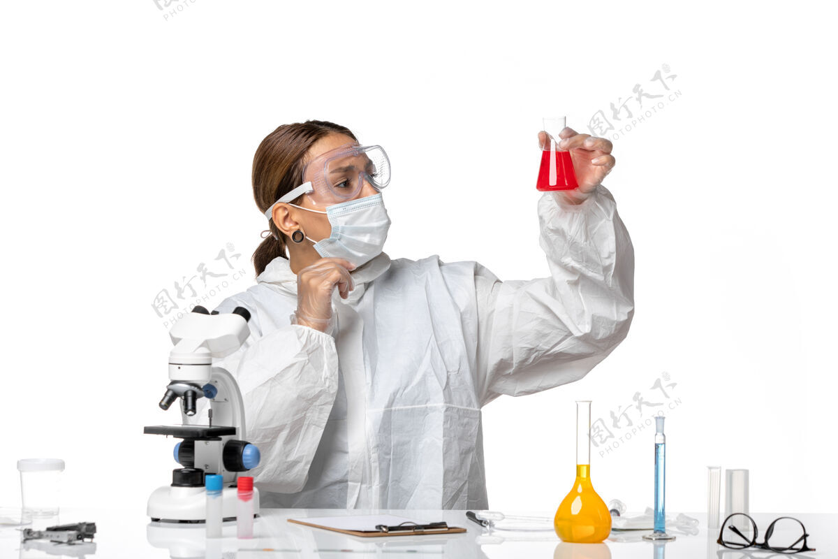 红色正面图：女医生穿着防护服 戴着口罩 拿着装有红色溶液的烧瓶 白色办公桌上有冠状病毒大流行烧瓶人药品