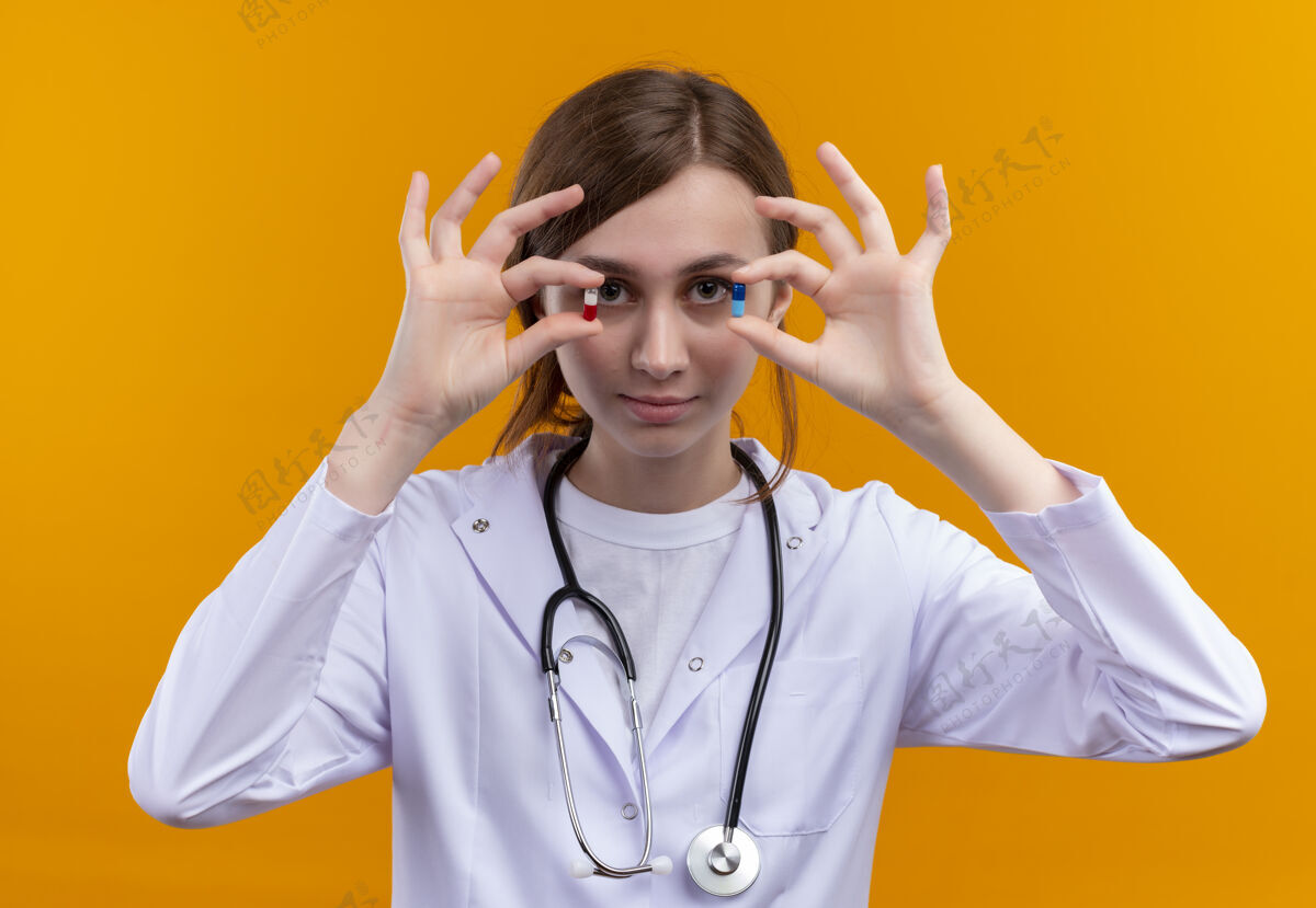 穿着自信的年轻女医生穿着医用长袍和听诊器在孤立的橙色空间展示医用胶囊胶囊展示听诊器