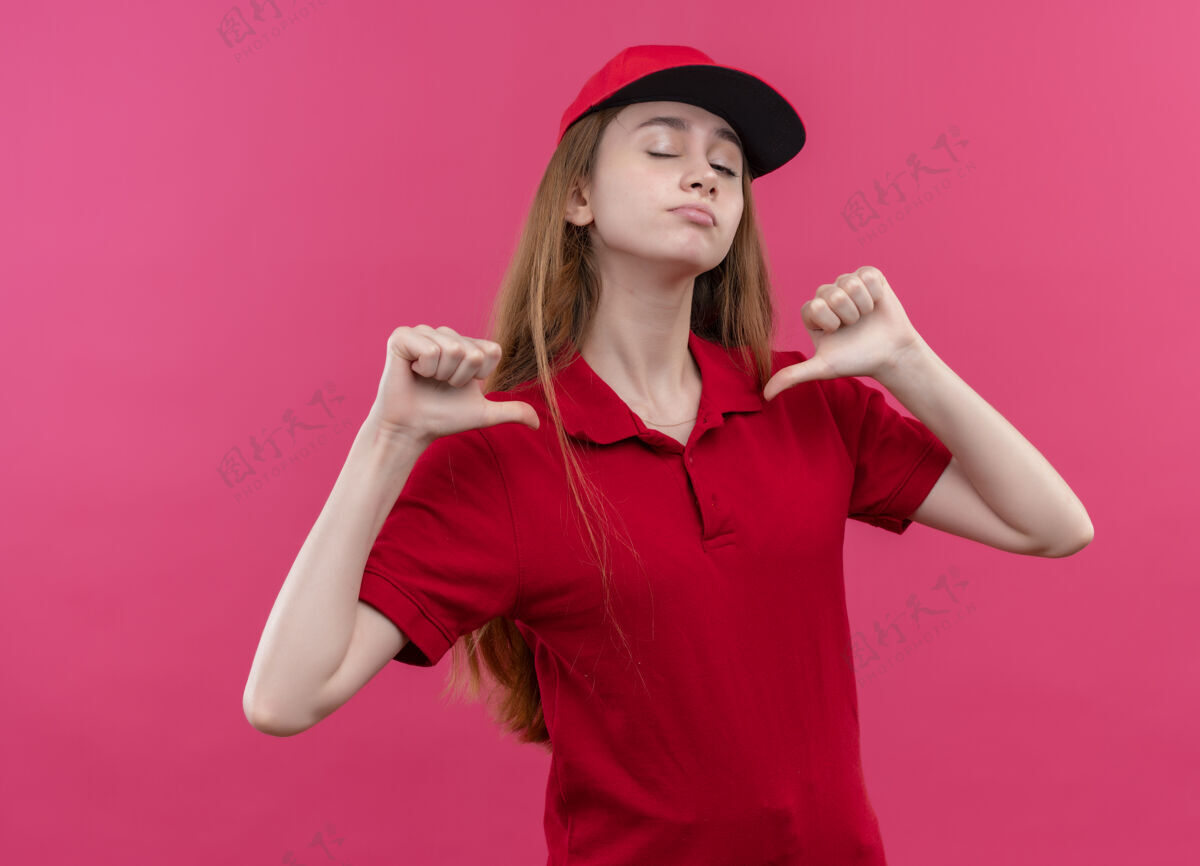 粉色穿着红色制服的自信的年轻送货女孩指着自己 在孤立的粉色空间里眨眼年轻制服女孩