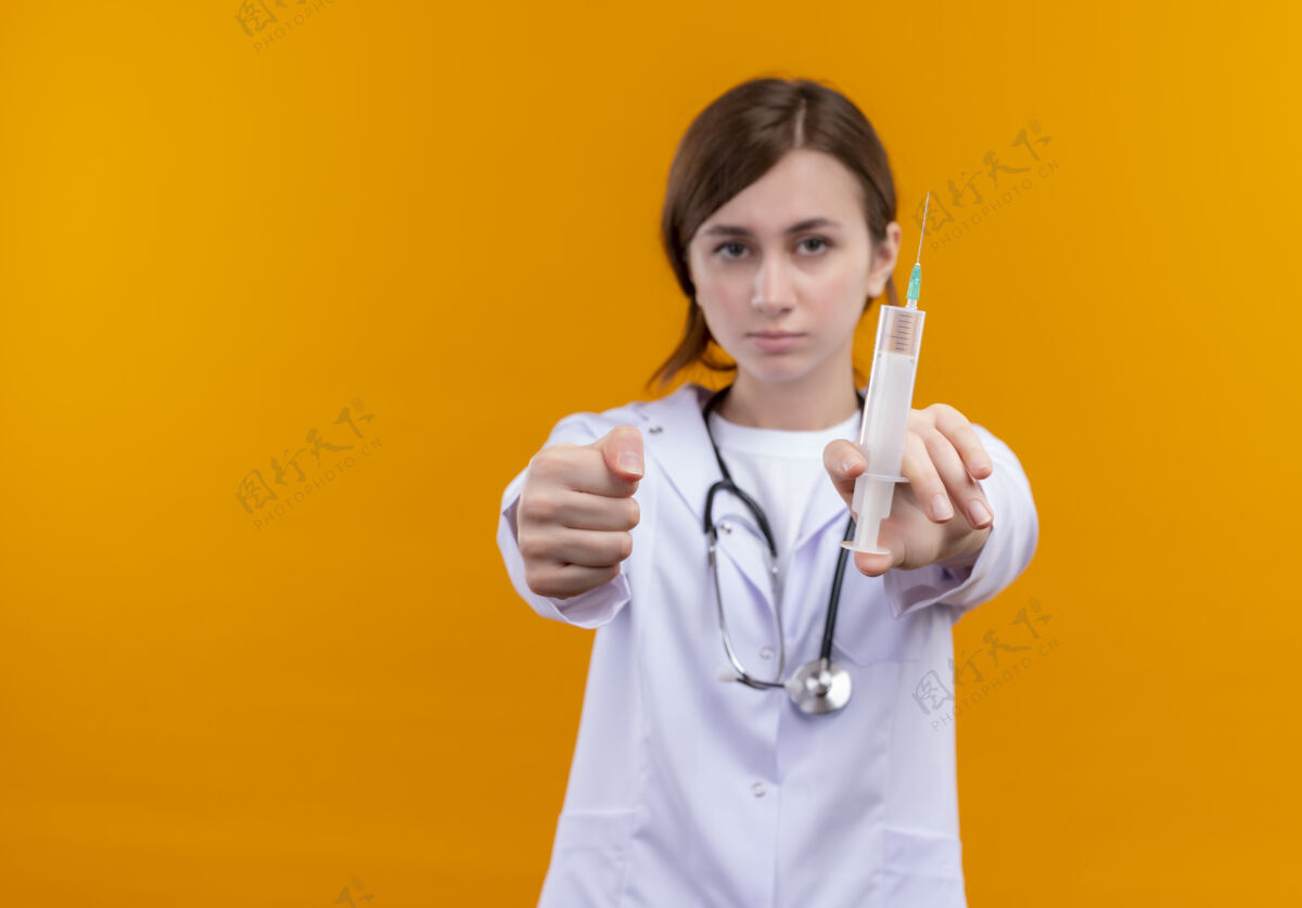 长袍自信的年轻女医生穿着医用长袍和听诊器伸展注射器和拳头在孤立的橙色空间与复印空间听诊器穿医生