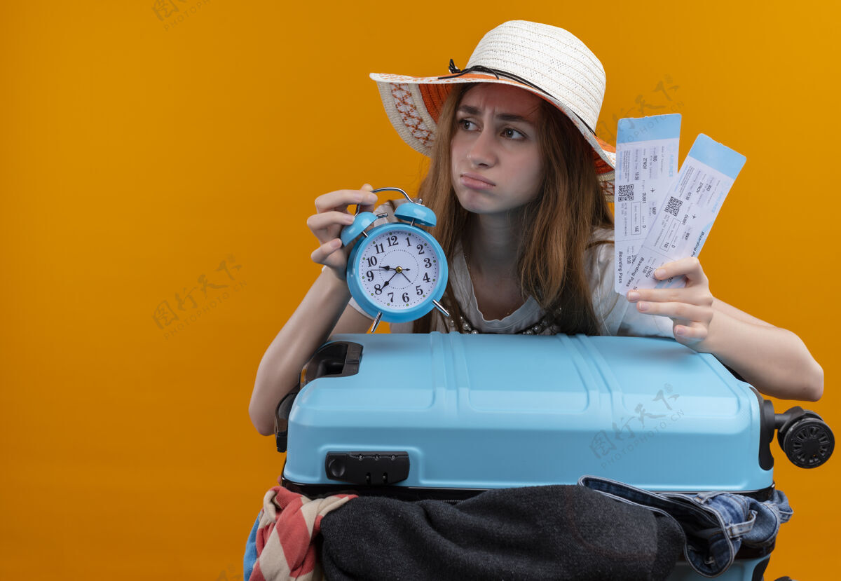橙色焦虑的年轻旅客女孩戴着帽子拿着飞机票和闹钟带着手提箱在孤立的橙色空间与复印空间女孩焦虑空间