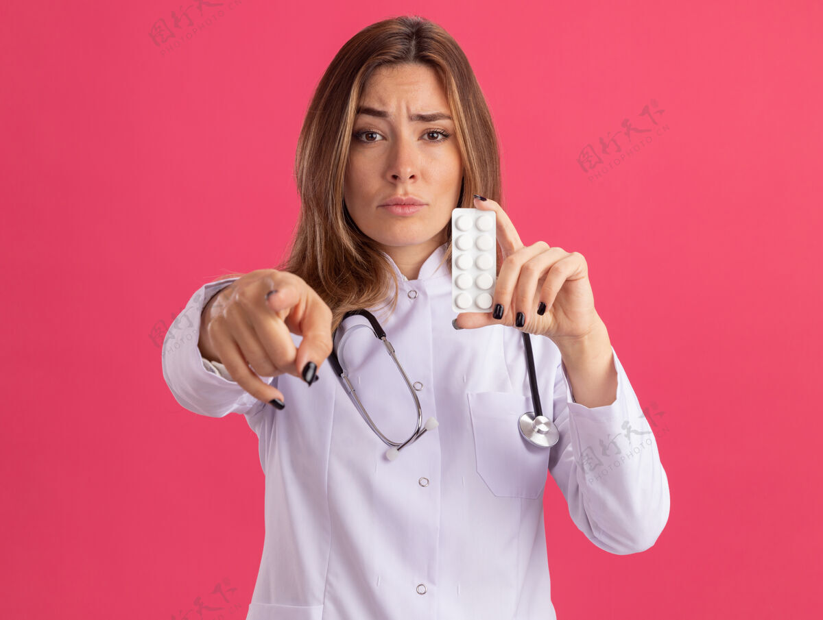 表情自信的年轻女医生穿着带听诊器的医用长袍 手持药片 站在隔离在粉红色墙上的前面女性长袍年轻人