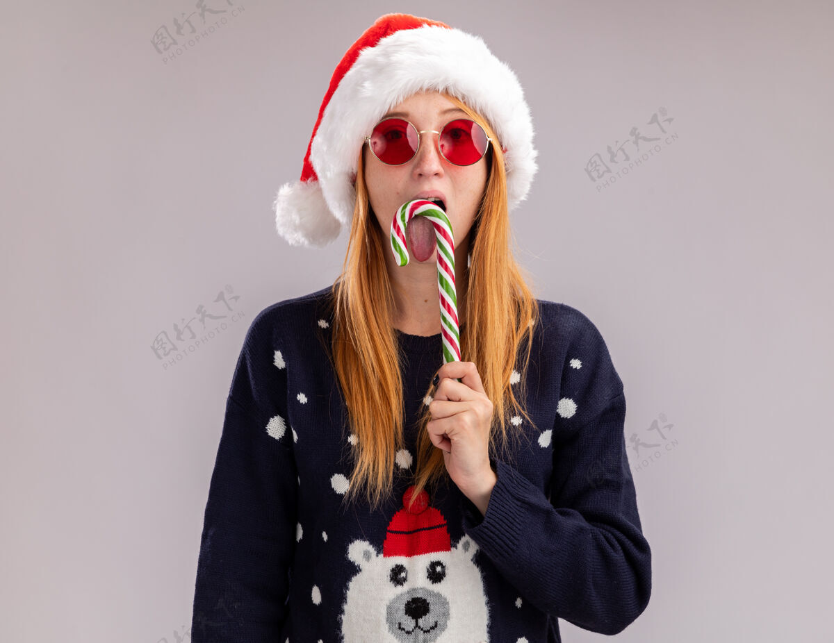 圣诞节看着镜头年轻漂亮的女孩戴着圣诞帽和眼镜拿着和舔着圣诞糖果隔离在白色背景上眼镜舔女孩