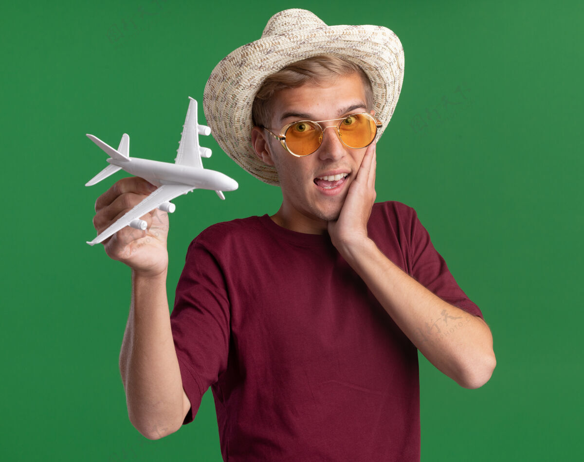 市民惊讶的年轻帅哥穿着红衬衫戴着眼镜和帽子拿着玩具飞机把手放在脸颊上隔离在绿色的墙上站着帅哥眼镜
