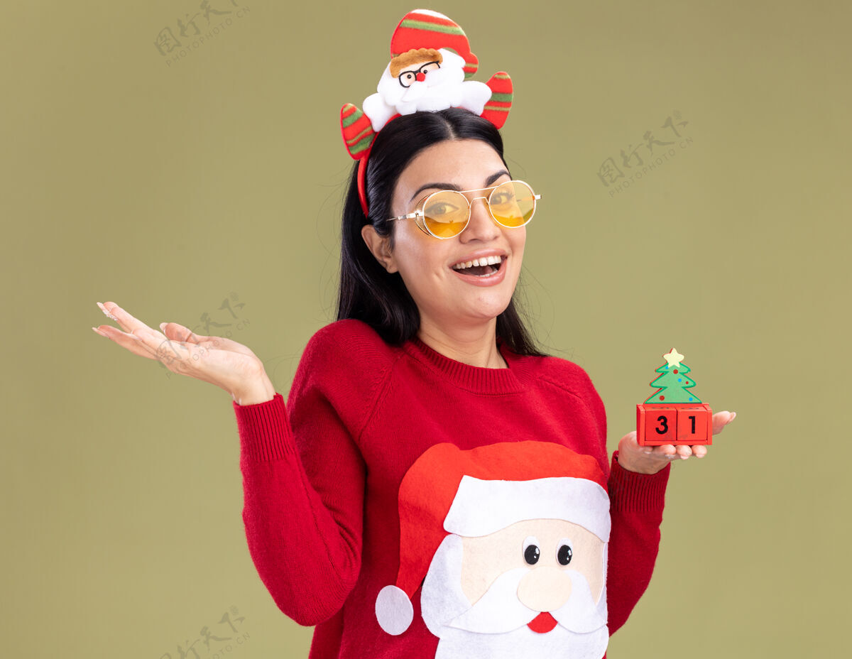 欢乐快乐的年轻白人女孩戴着圣诞老人的头带和毛衣 戴着眼镜 手里拿着圣诞树玩具 手里拿着日期 看着相机 在橄榄绿的背景上 一只空手孤立着头带圣诞老人手