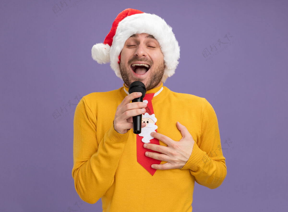 麦克风快乐的年轻白种人戴着圣诞帽打着领带拿着麦克风闭着眼睛唱歌手放在胸前紫色背景上隔离手年轻领带