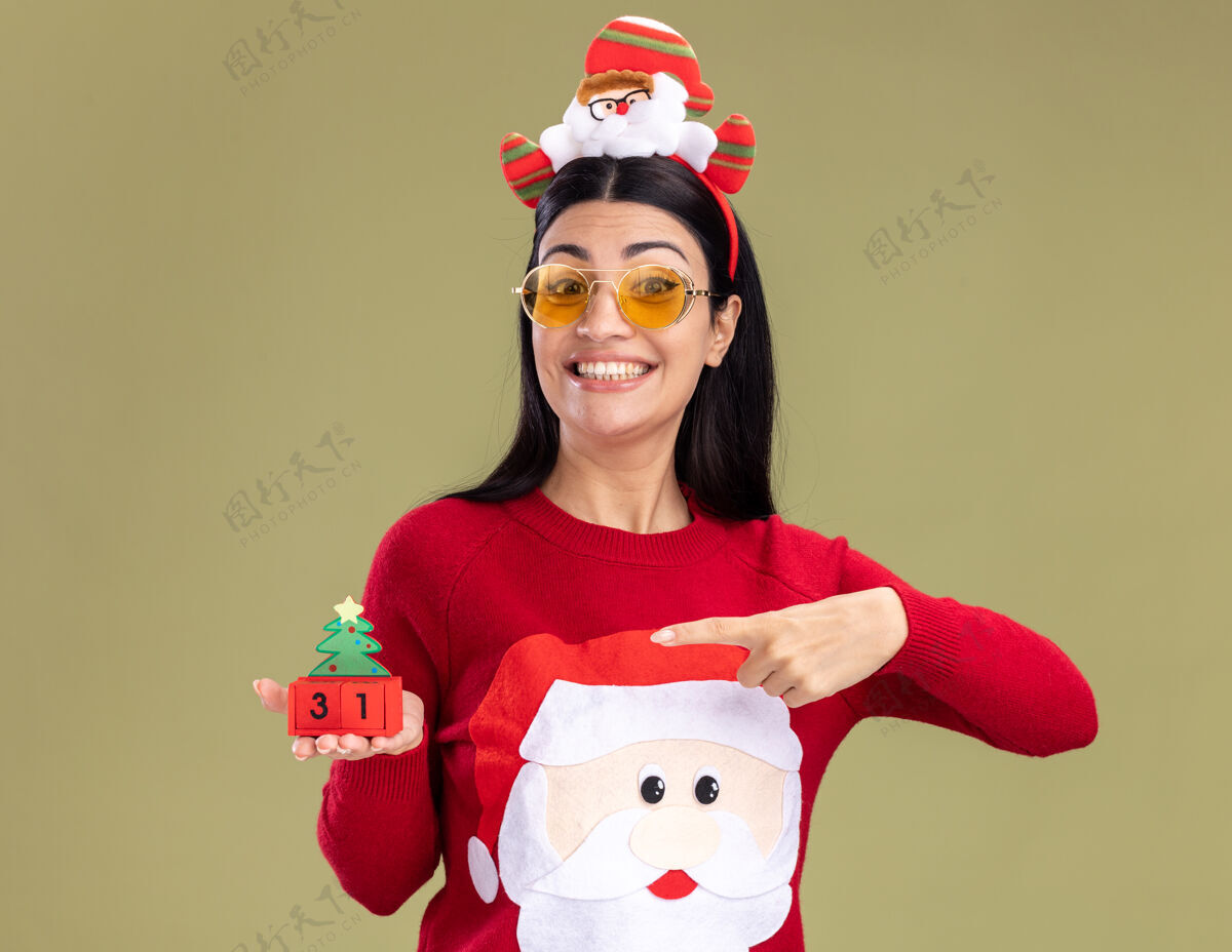 毛衣快乐的年轻白人女孩戴着圣诞老人的头带和毛衣 戴着眼镜 拿着并指着圣诞树玩具和日期 看着橄榄绿背景上孤立的相机圣诞指着圣诞快乐