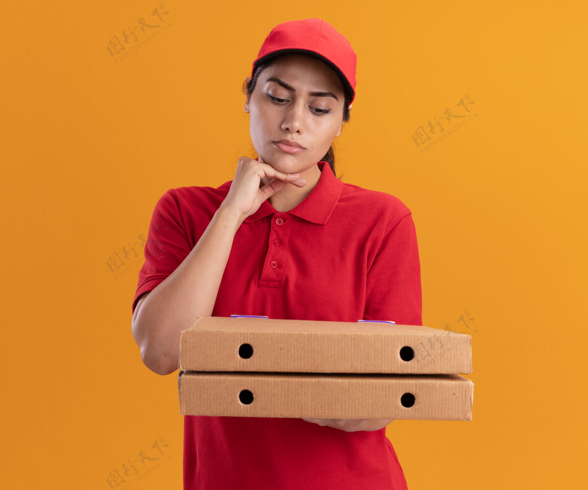 下巴想着穿着制服戴着帽子的年轻送货女孩 看着披萨盒把手放在橘色墙上的下巴下面公民人披萨