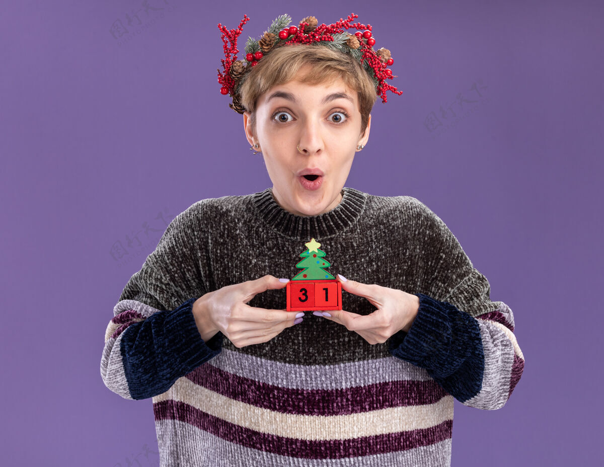 圣诞快乐令人印象深刻的年轻漂亮女孩戴着圣诞花环拿着圣诞树玩具和日期看着相机隔离在紫色的背景紫色年轻圣诞树