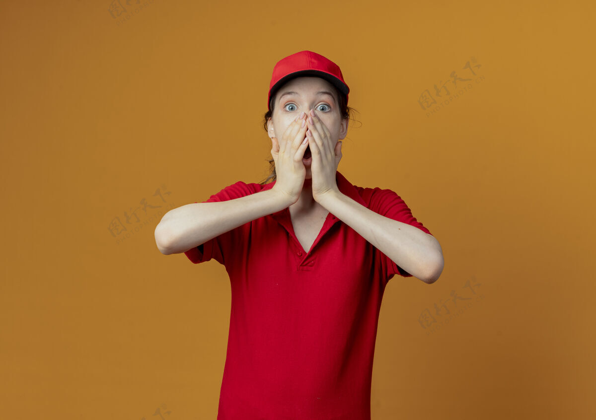 交货惊讶的年轻漂亮的送货女孩穿着红色制服 戴着帽子 手放在嘴上 看着橙色背景上的相机 还有复印空间帽子惊喜红色