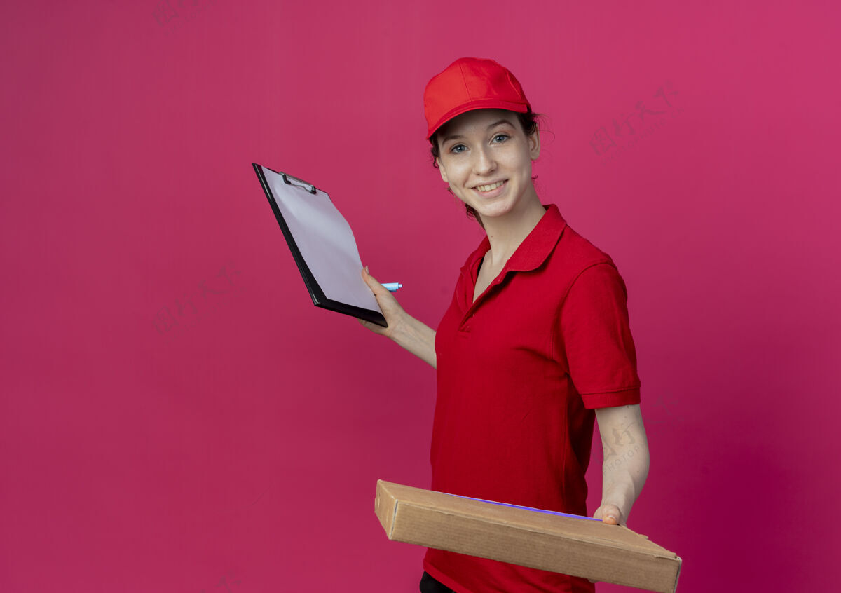 背景微笑着的年轻漂亮的送货女孩 穿着红色制服 戴着帽子 手里拿着披萨包和剪贴板 笔被隔离在深红色的背景上 还有复印空间披萨深红色女孩