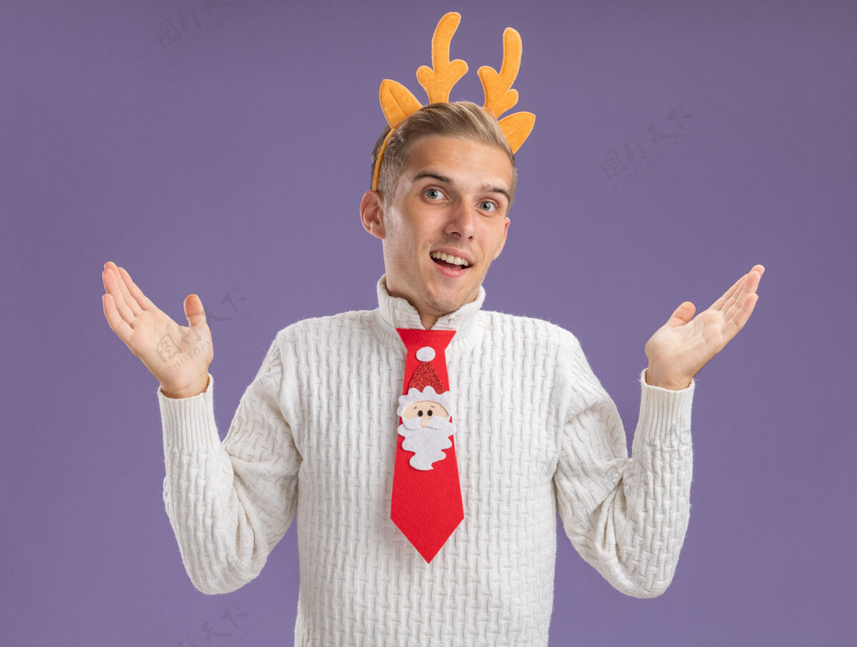 头带令人印象深刻的年轻帅哥戴着驯鹿鹿角头带和圣诞老人领带看着相机显示空手孤立在紫色的背景年轻空领带