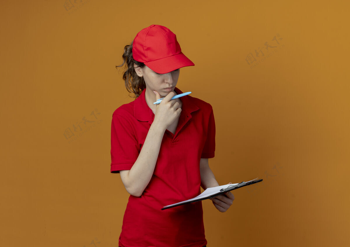 剪贴板体贴的年轻漂亮的送货女孩 穿着红色制服 戴着帽子 触摸着下巴 拿着剪贴板和笔 看着孤立在橙色背景上的剪贴板 还有复印空间年轻橙色思想