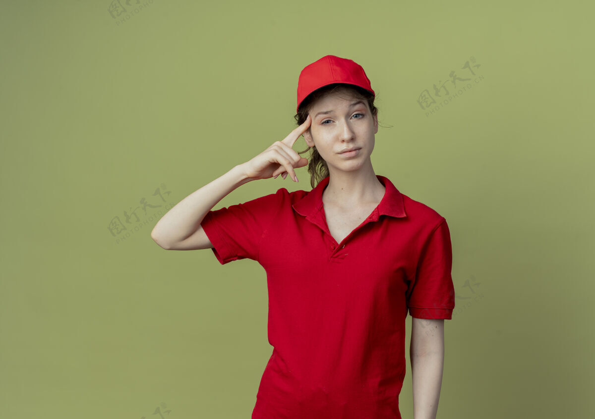 年轻自信的年轻漂亮的送货女孩 穿着红色制服 戴着帽子 手指放在太阳穴上 在橄榄绿的背景上 留有复印空间太阳穴女孩漂亮