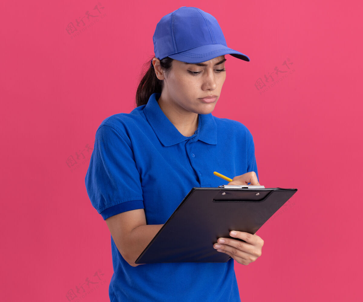 年轻自信的年轻送货女孩穿着制服 戴着帽子 在粉色墙上的剪贴板上写着什么女孩制服表情