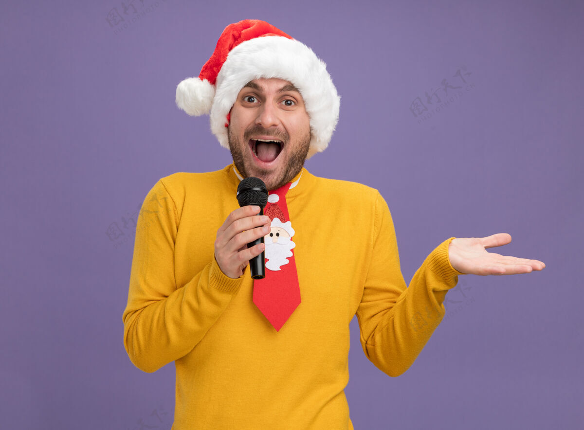 男人令人印象深刻的年轻白种人戴着圣诞帽 打着领带 拿着麦克风 看着镜头 紫色背景上孤立的空手年轻帽子麦克风