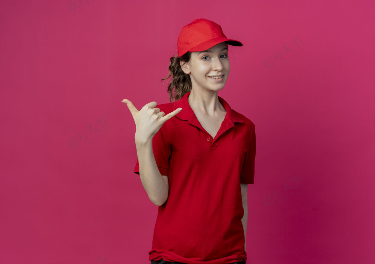 制服微笑着的年轻漂亮的送货女孩 穿着红色制服 戴着帽子 在深红色的背景上做着一个松散的姿势深红色松散红色