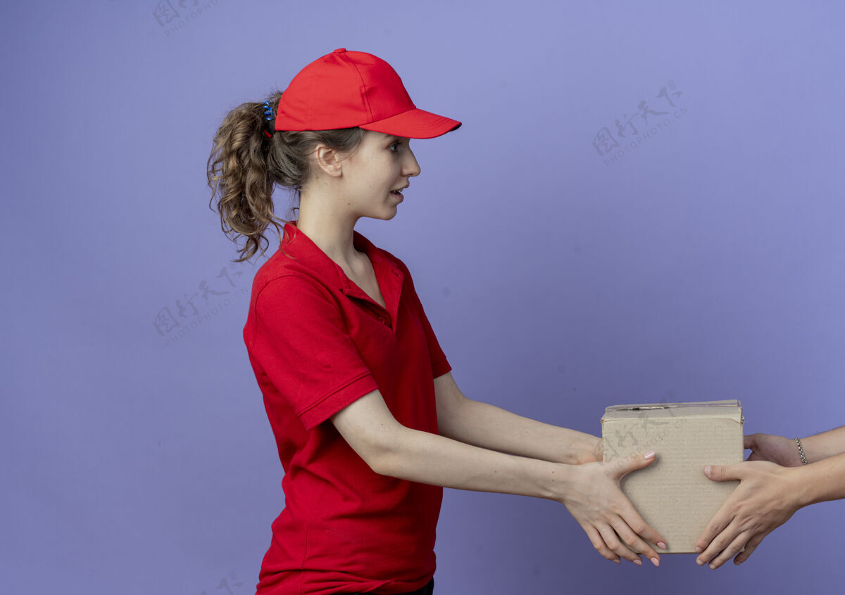 客户令人印象深刻的年轻漂亮的送货女孩穿着红色制服 戴着帽子站在侧视图给客户纸箱在紫色背景与复制空间隔离漂亮交货纸箱