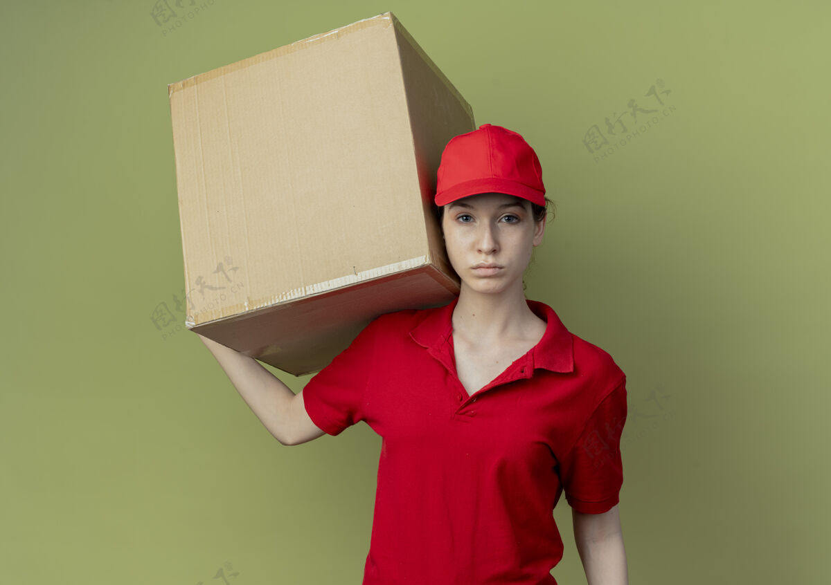 漂亮年轻漂亮的送货女孩 穿着红色制服 戴着帽子 肩上扛着纸箱 看着橄榄绿背景上的相机 还有复印空间橄榄制服年轻
