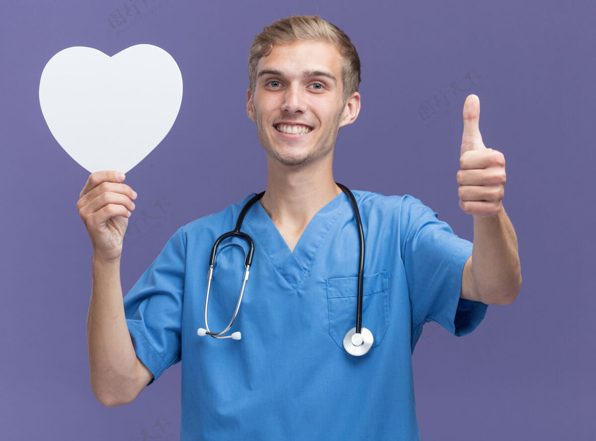 人微笑的年轻男医生穿着医生制服 手持听诊器 手持心形盒子 大拇指向上竖立在蓝色的墙上盒子穿着人
