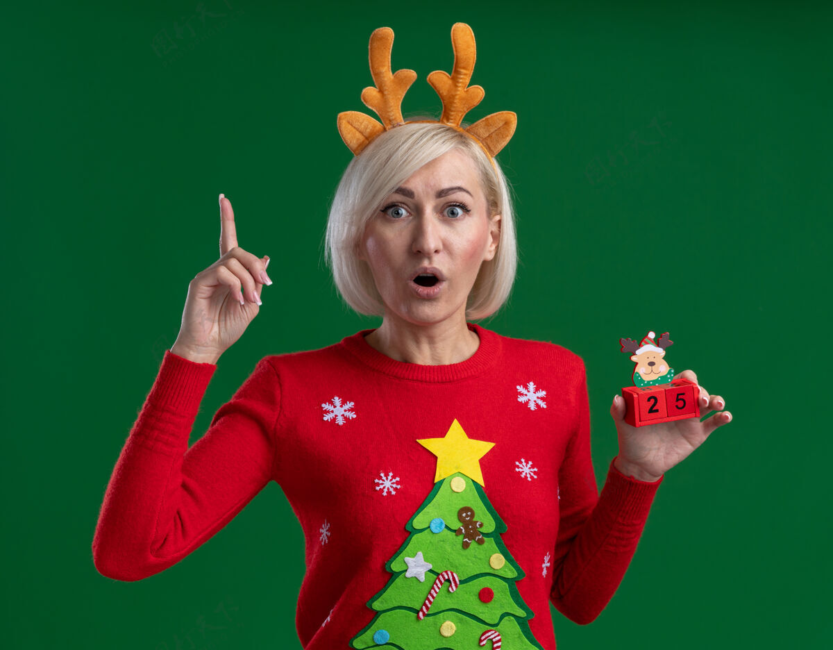 指向令人印象深刻的中年金发女人戴着圣诞驯鹿鹿角头带和圣诞毛衣拿着圣诞驯鹿玩具和日期看着相机指向上孤立的绿色背景举行向上鹿角