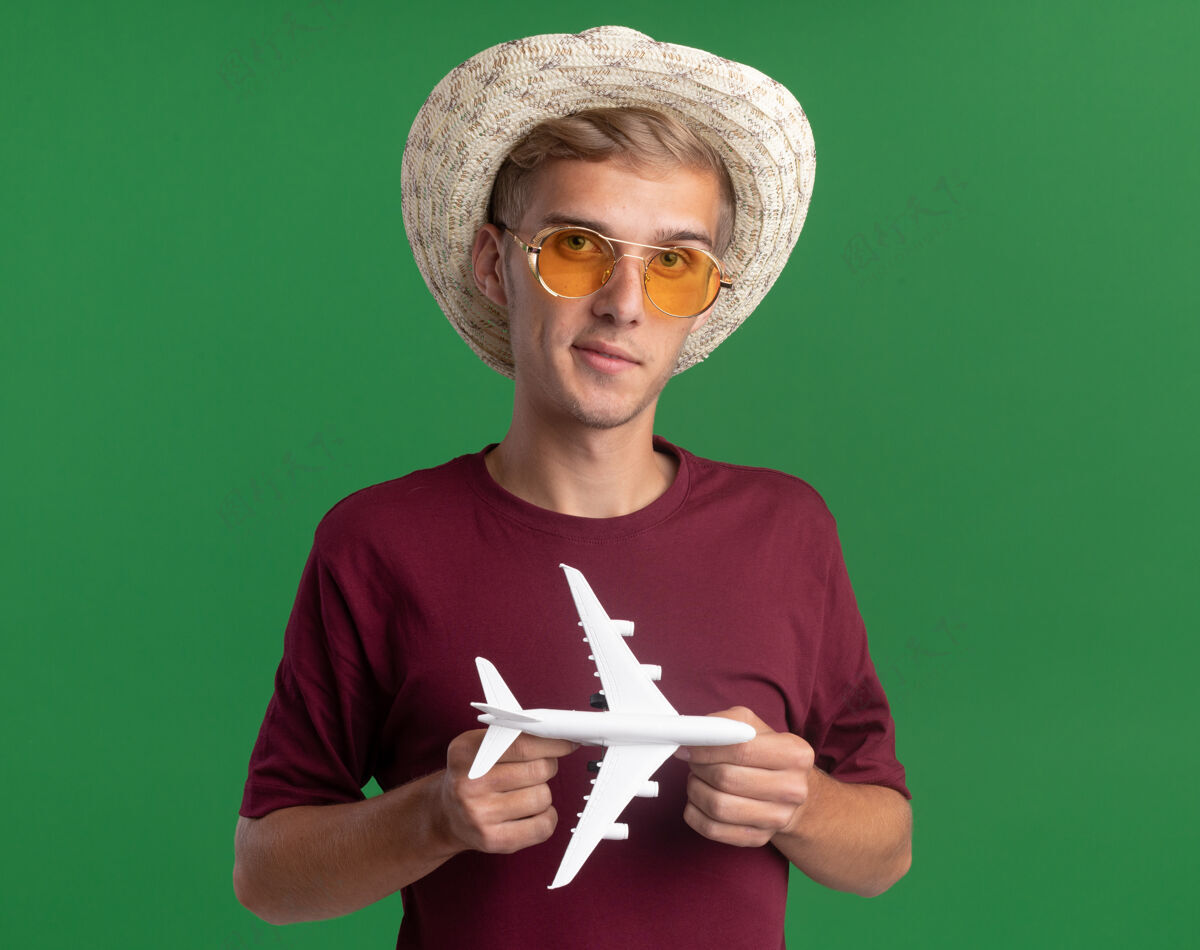 感觉高兴的年轻帅哥穿着红衬衫戴着眼镜和帽子拿着玩具飞机隔离在绿色的墙上市民人年轻人
