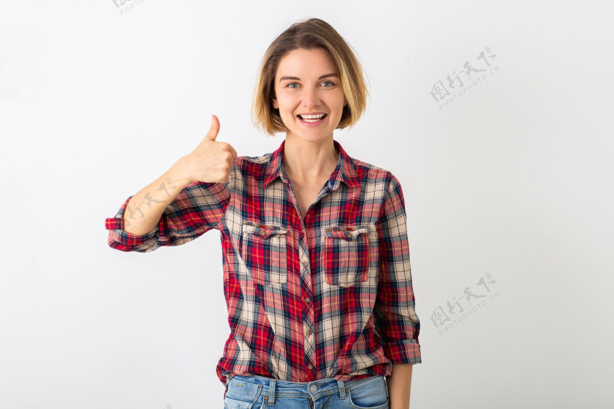竖起大拇指穿着格子衬衫的年轻有趣的情绪化女人在白色的工作室墙上摆出孤立的姿势 展示拇指向上的姿势肖像随意姿势