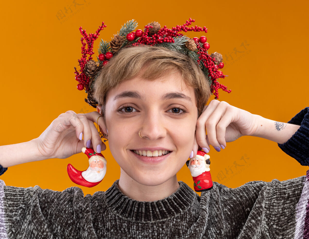 圣诞老人特写镜头：微笑的年轻漂亮女孩戴着圣诞花环 耳朵上挂着圣诞老人的圣诞饰品 看着隔离在橙色背景上的相机头圣诞老人圣诞
