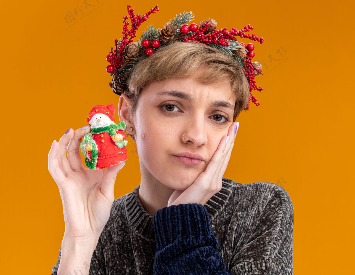 年轻困惑的年轻漂亮女孩戴着圣诞花环手持小圣诞雪人雕像手放在脸上看着隔离在橙色背景下的相机圣诞快乐头手