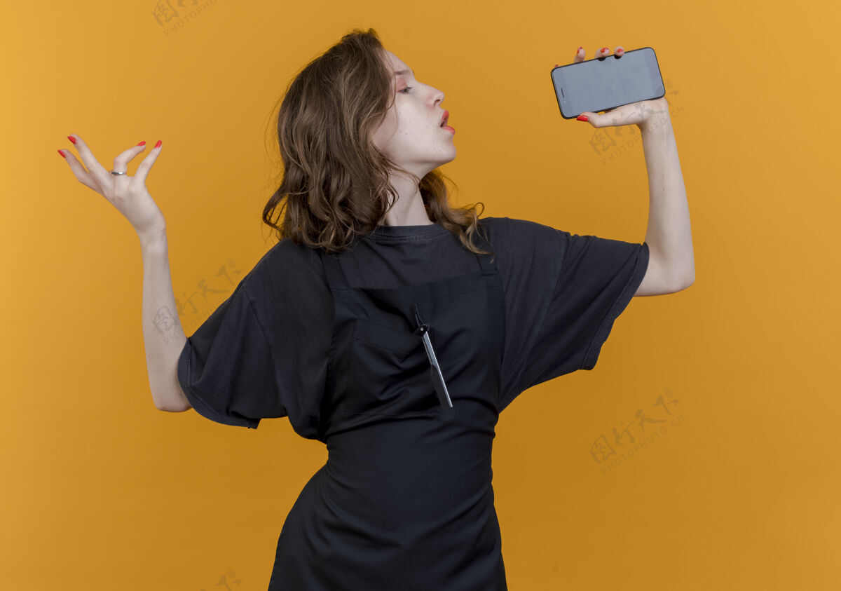 年轻年轻的斯拉夫女理发师穿着制服 用手机作为麦克风唱歌 手在橙色背景下与空气隔绝手橙色保持