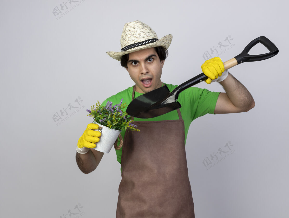 年轻穿着制服的年轻男园丁惊讶地戴着园艺帽和手套 拿着铲子指着花盆里的花举行穿黑桃