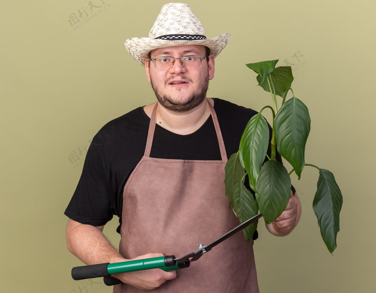 花园高兴的年轻男园丁戴着园艺帽拿着修剪器隔离在橄榄绿墙上的植物帽子姿势脸