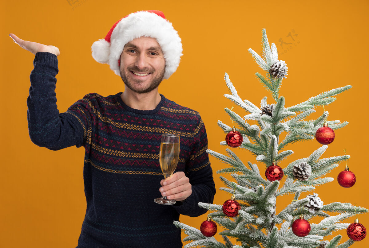 男人快乐的白人年轻人戴着圣诞帽 站在装饰好的圣诞树旁 手里拿着一杯香槟 看着摄像机 在橙色背景下 一只空手孤立着圣诞站着年轻