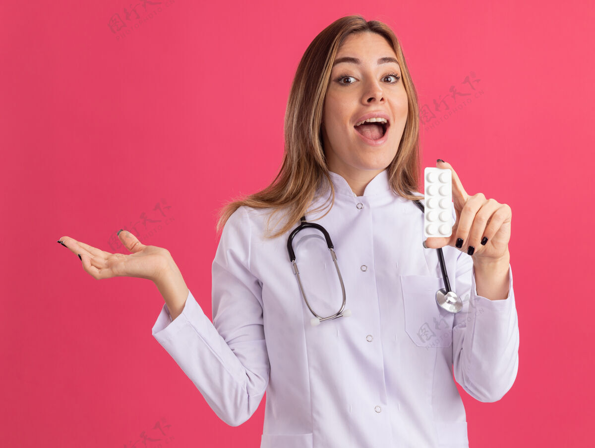 人兴奋的年轻女医生 穿着医用长袍 手持听诊器 手被隔离在粉红色的墙上 手里拿着药片女性衣服人