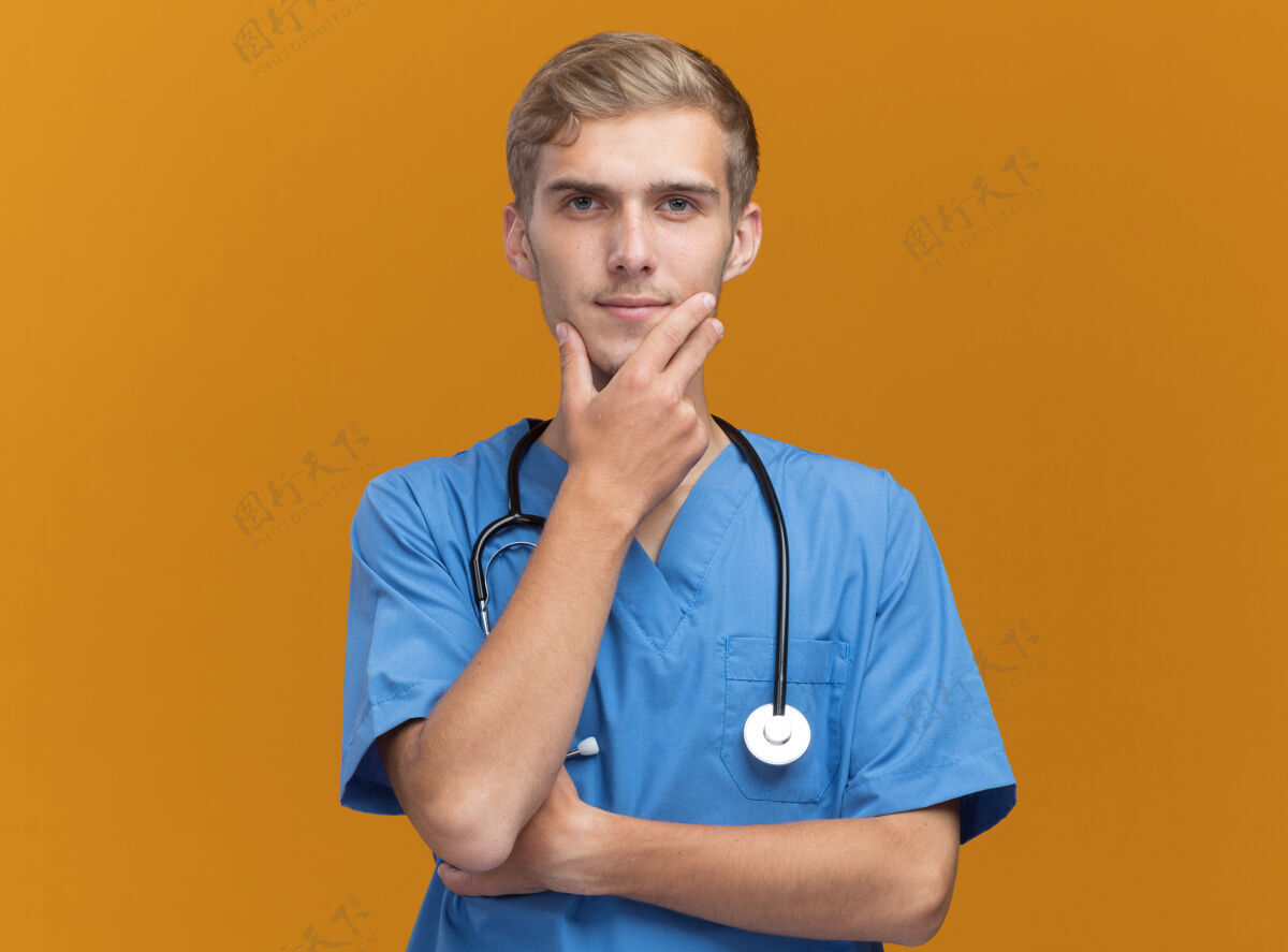 衣服自信的年轻男医生穿着医生制服 手持听诊器 把下巴孤立地抓在橙色的墙上站立表情年轻人