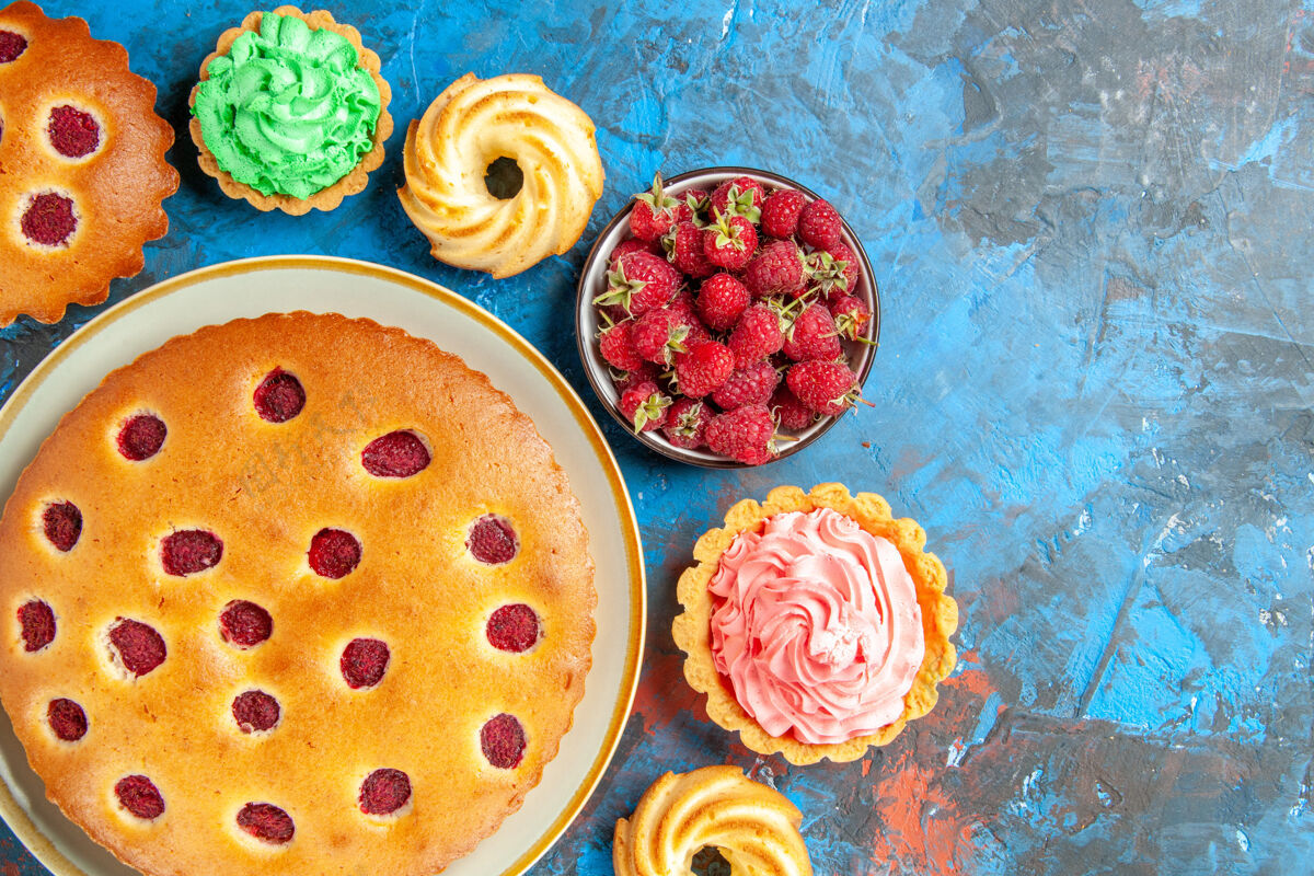 椭圆形椭圆形盘子上覆盆子蛋糕的俯视图 四周是饼干 小馅饼和蓝色表面有浆果的碗正餐碗美味