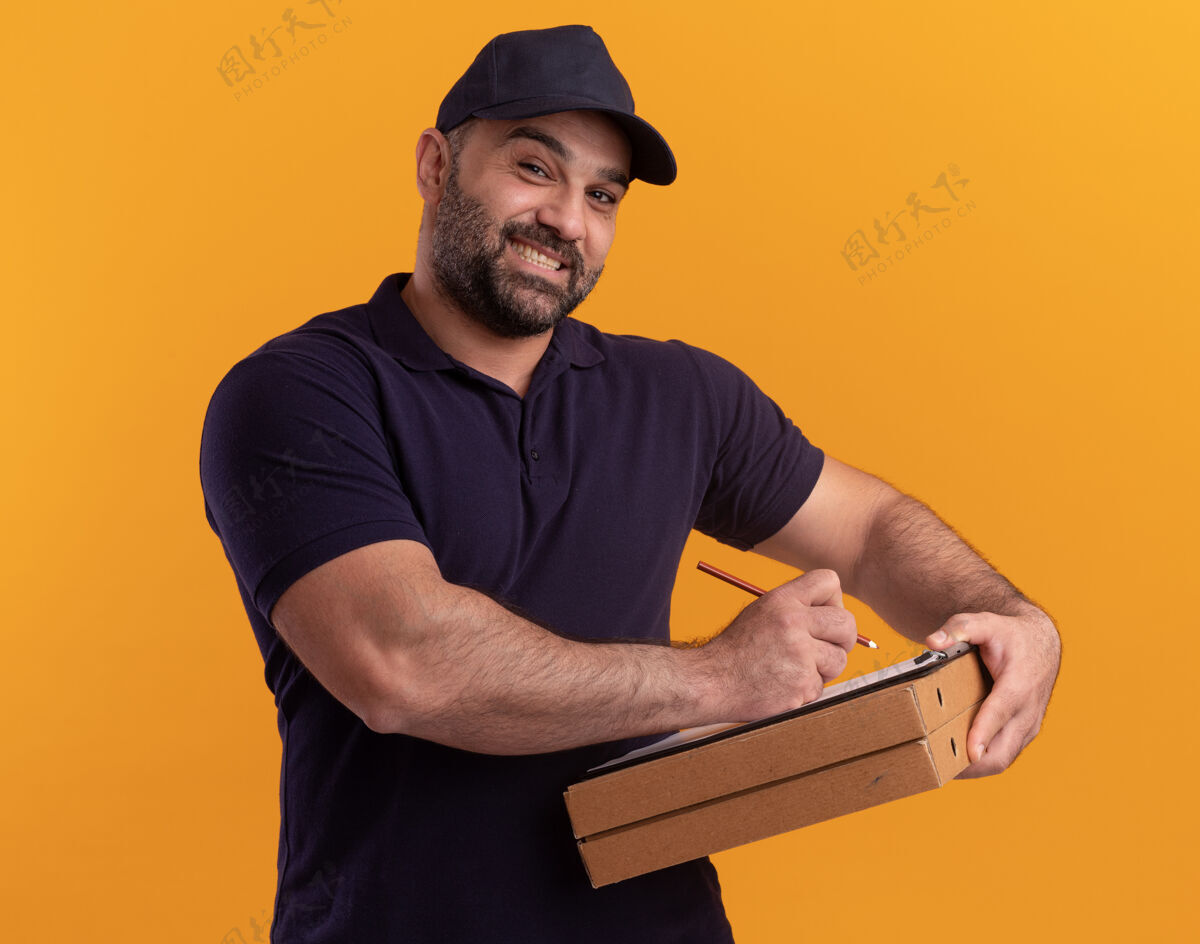 站微笑着的中年送货员 穿着制服 戴着帽子 在黄色墙上的比萨饼盒上的剪贴板上写着什么书写微笑衣服