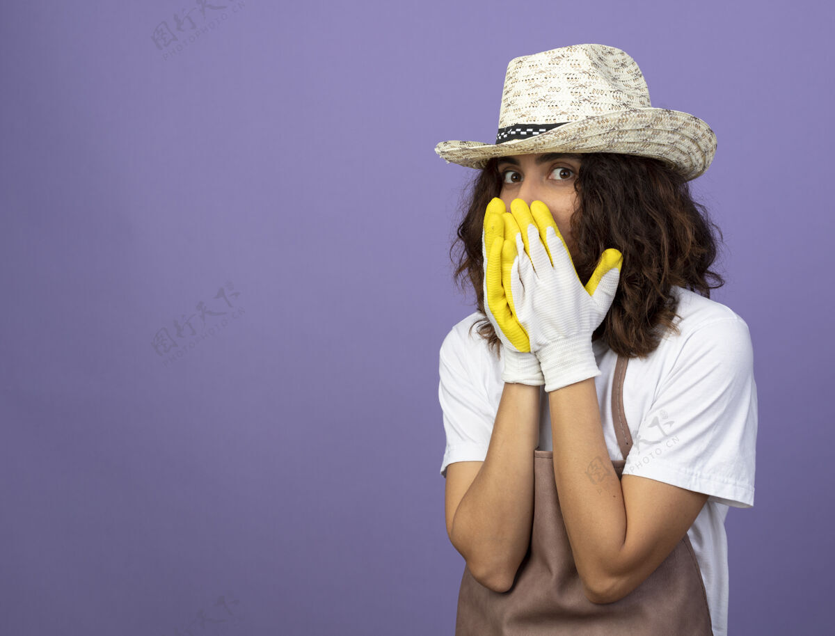 成人惊讶的年轻女园丁穿着制服戴着园艺帽和手套用手捂住脸手套紫色花园