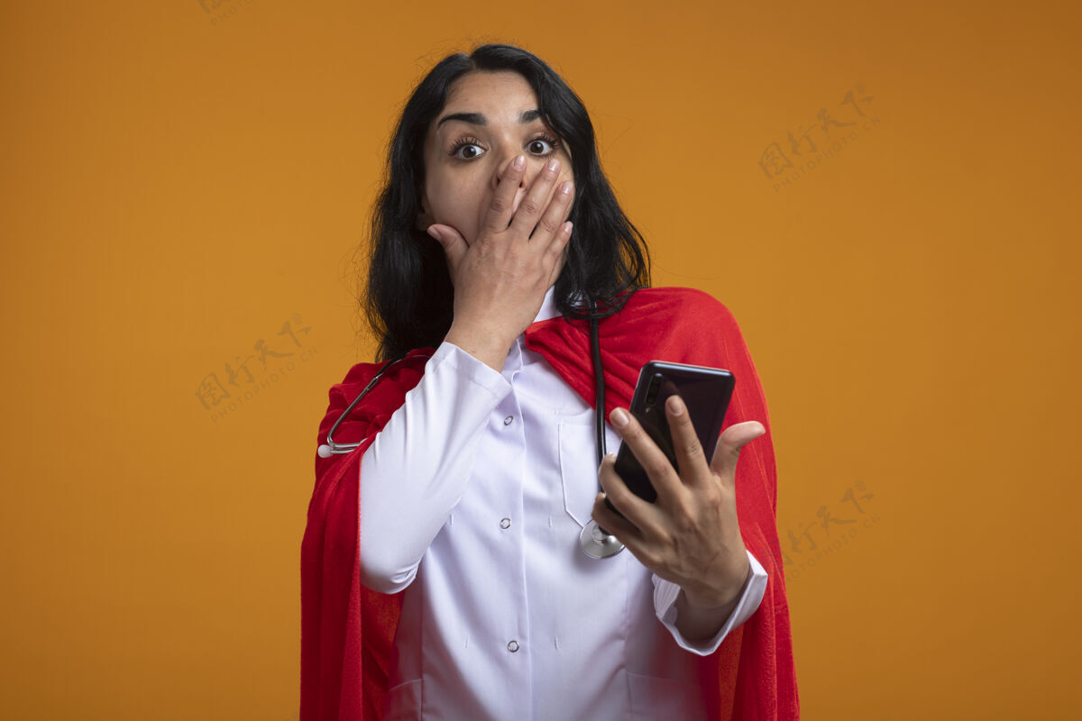 超级英雄被吓坏了的年轻超级英雄女孩 穿着医用长袍 手持听诊器 嘴上盖着电话 手被隔离在橙色的墙上电话年轻女孩