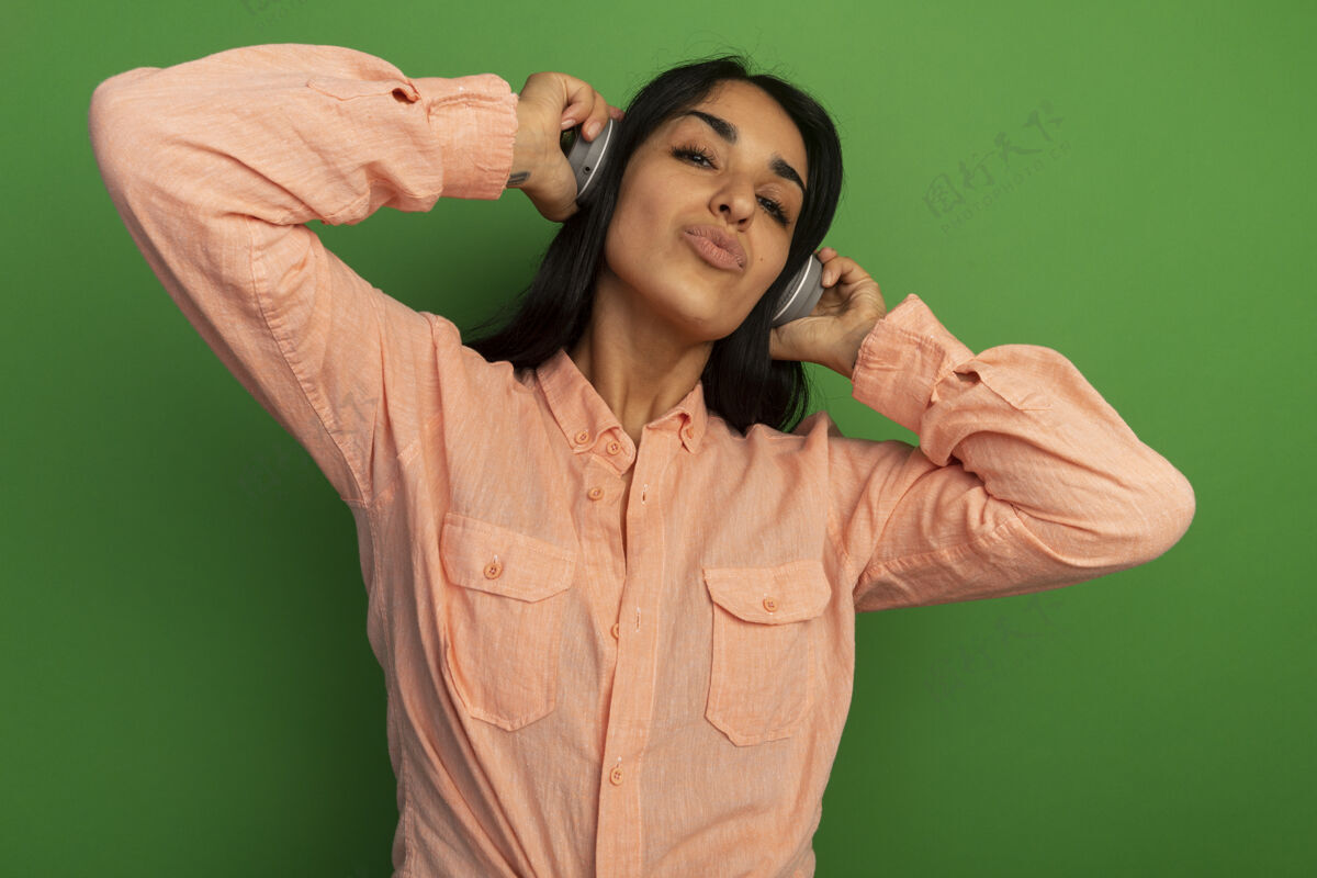 耳机高兴的年轻漂亮的女孩穿着粉红色的t恤和耳机隔离在绿色的墙壁上女孩请戴上年轻人