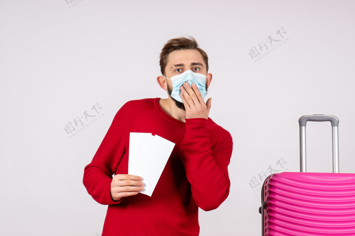 颜色正面图：戴着无菌口罩的年轻男性手持机票在白墙上航行covid-旅行度假情感病毒航班颜色微笑肖像情绪