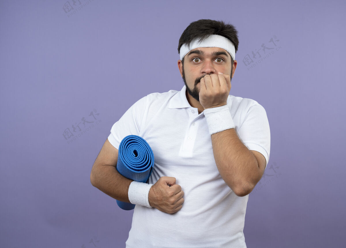 抱着戴着头带和腕带的关心运动的年轻人拿着瑜伽垫 手放在嘴上 隔离在绿色的墙壁上 有复制空间垫子腕带头带