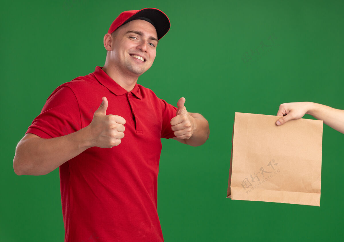 年轻人微笑着的年轻送货员穿着制服 戴着帽子 把纸食品包递给客户 在绿色的墙上竖起大拇指穿着顾客给予