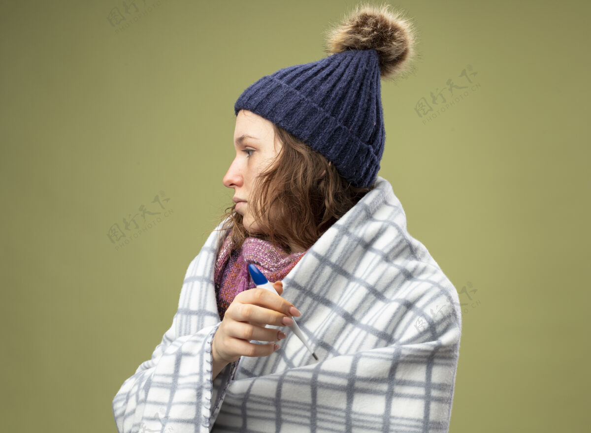 疾病站在侧视图年轻的生病的女孩穿着白色长袍和冬季帽子围巾包裹在格子布举行温度计隔离在橄榄绿帽子橄榄色围巾